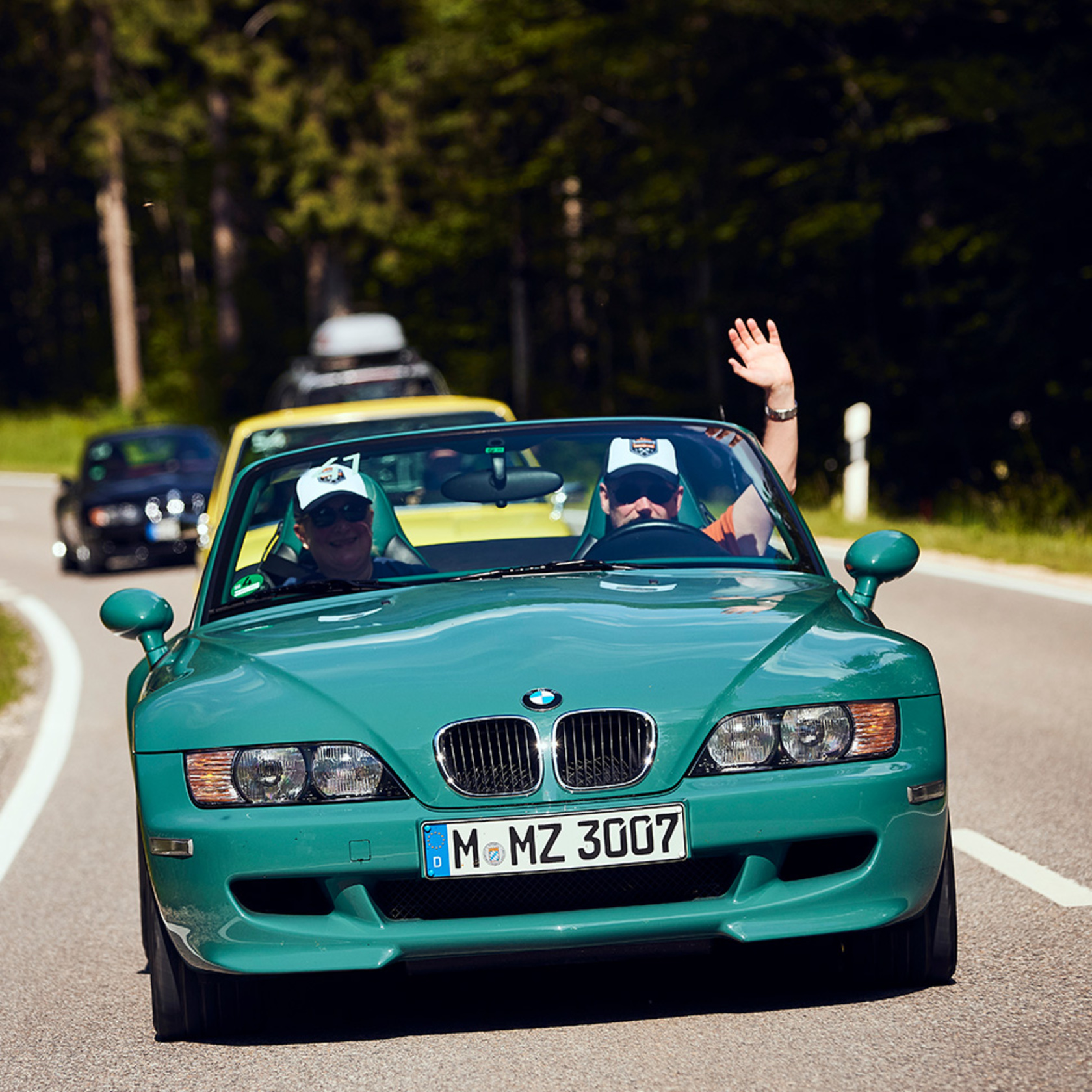 zwei Männer mit weißen Schirmmützen fahren in einem türkisfarbenen BMW Cabrio die Straße entlang und winken in die Kamera