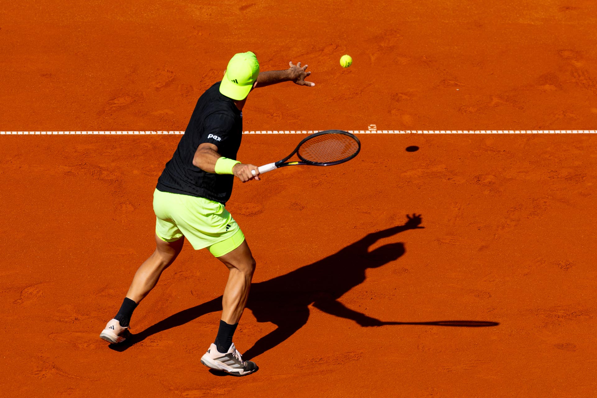Ein Tennisspieler in schwarzem T-shirt, sowie neongrüner Hose und Schirmmütze schlägt einen Ball